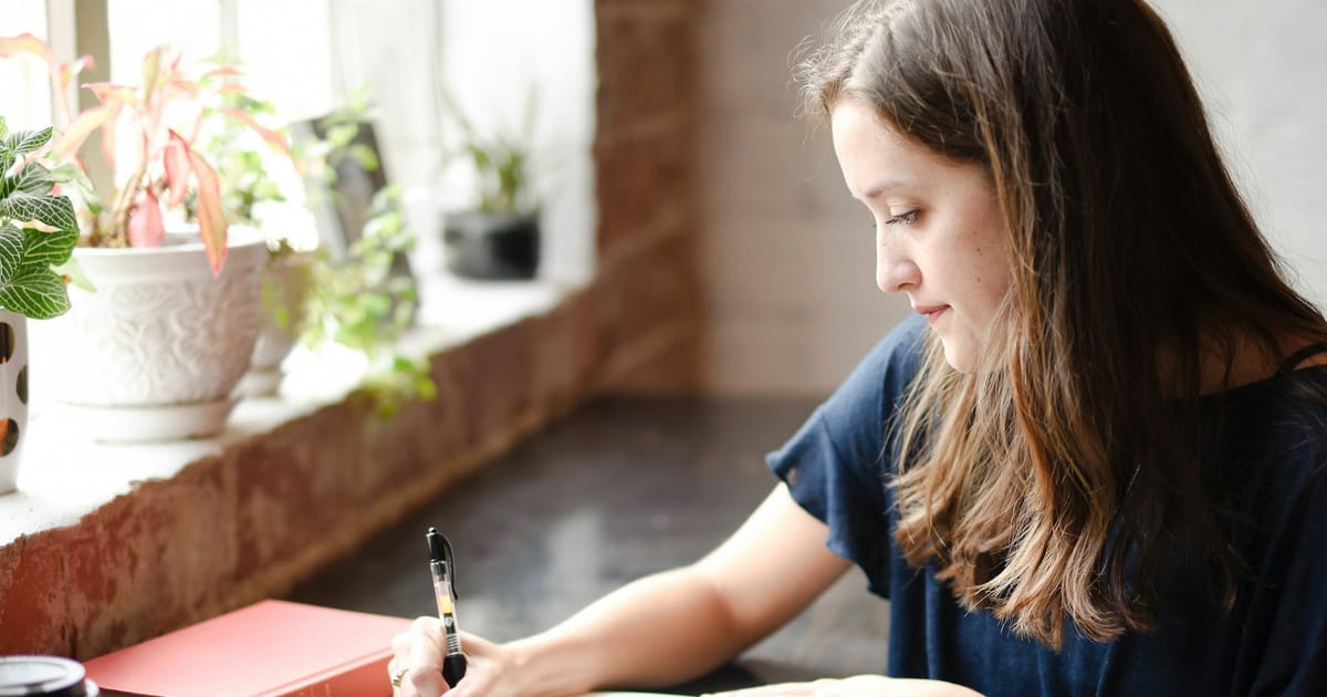Una mujer escribe en un cuaderno junto a una ventana con luz natural.