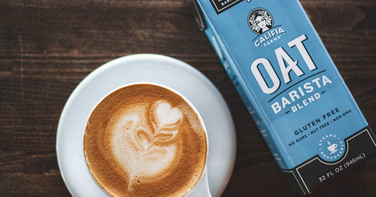 Una taza de café con leche junto a una caja de leche de avena para barista.