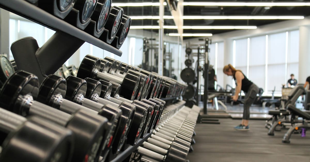 Una mujer entrena en un gimnasio amplio, detrás de un estante lleno de mancuernas.